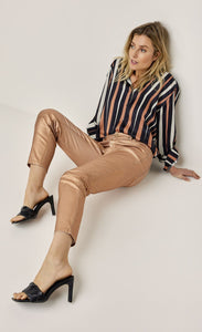 Front full body view of a woman sitting, wearing a black, pink, and white striped shirt and the summum skinny foil coated trousers. These trousers are rose gold colored.
