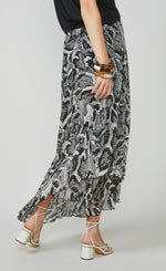Load image into Gallery viewer, Back bottom half view of a woman wearing the summum printed maxi skirt. This skirt has a black and white snake skin print with a black waistband. The skirt has an asymmetrical hem and a flowy look.
