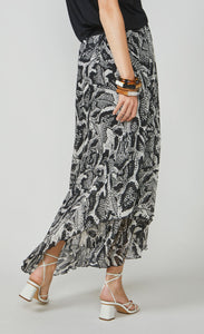Back bottom half view of a woman wearing the summum printed maxi skirt. This skirt has a black and white snake skin print with a black waistband. The skirt has an asymmetrical hem and a flowy look.