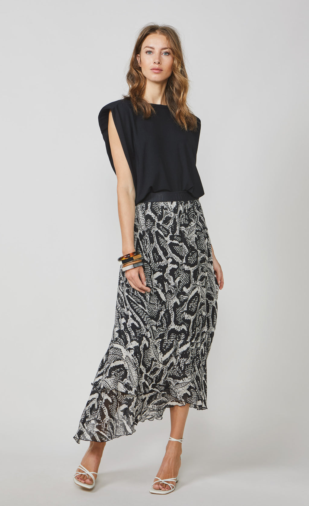 Front full body view of a woman wearing the summum black shoulder pad top and the summum printed maxi skirt. This skirt has a black and white snake skin print with a black waistband. The skirt has an asymmetrical hem and a flowy look.