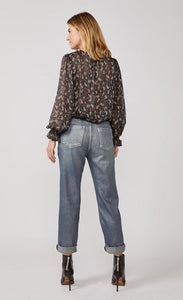 Back Full body view of a woman wearing a black top with print and the summum straight blue daze denim pants. These jeans are blue-grey with a shimmer. They have a cropped and cuffed bottom, pockets, and a tie belt.