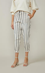 Load image into Gallery viewer, Front bottom half view of a woman wearing a white top and the summum striped cotton trouser. This pant is ivory colored with black stripes. It is wider around the hips and tapers in above the ankles where it ends. 
