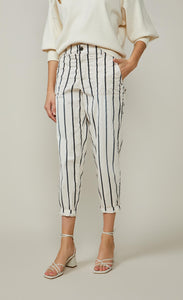 Front bottom half view of a woman wearing a white top and the summum striped cotton trouser. This pant is ivory colored with black stripes. It is wider around the hips and tapers in above the ankles where it ends. 