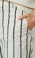 Load image into Gallery viewer, Front close up view of a woman wearing the summum striped cotton trouser. This pant is ivory colored with black stripes. This image shows off the side pocket.
