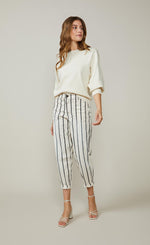 Load image into Gallery viewer, Front full body view of a woman wearing a white top and the summum striped cotton trouser. This pant is ivory colored with black stripes. It is wider around the hips and tapers in above the ankles where it ends. 
