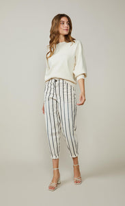 Front full body view of a woman wearing a white top and the summum striped cotton trouser. This pant is ivory colored with black stripes. It is wider around the hips and tapers in above the ankles where it ends. 