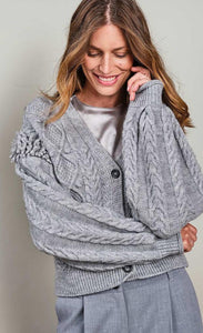 Front top half view of a woman wearing the summum balloon sleeve cardigan in the color fog/grey. This sweater has a cable knit pattern, a button down front, and fringe on the balloon sleeves.