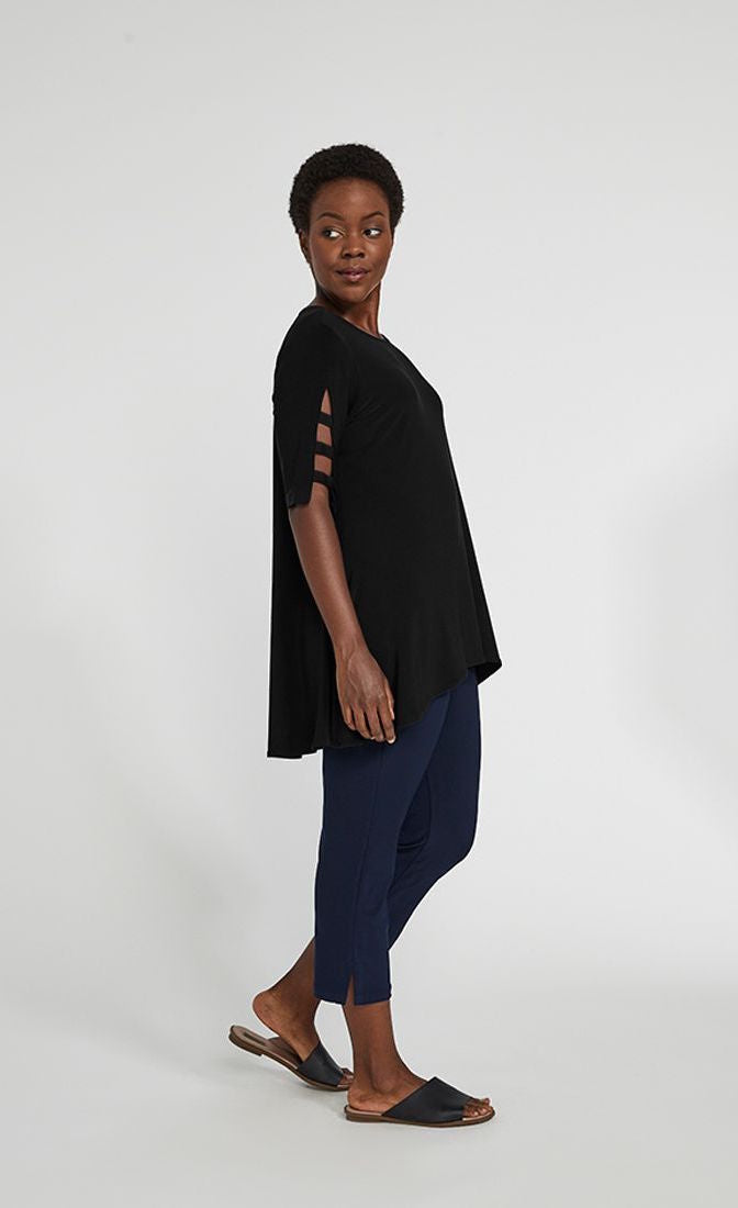 Left side full body view of a woman wearing blue pants and the sympli capture tunic in black. This top has short sleeves with striped slits, a round neck, and an arched high-low hem.