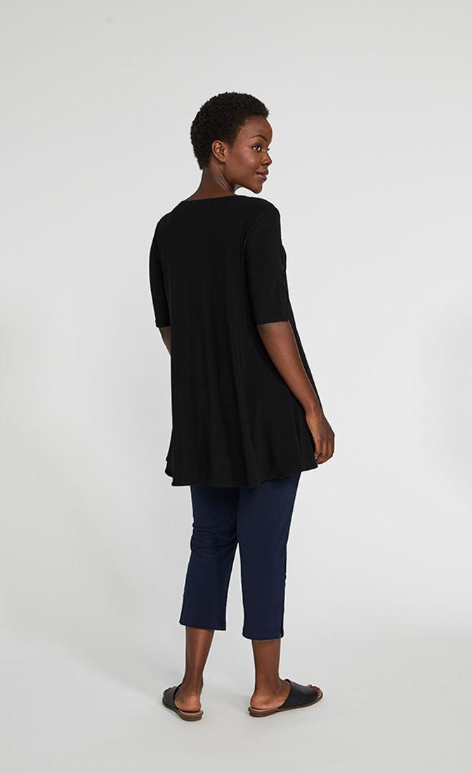 Back full body view of a woman wearing blue pants and the sympli capture tunic in black. This top has short sleeves with striped slits and an arched high-low hem.