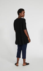 Load image into Gallery viewer, Back full body view of a woman wearing blue pants and the sympli capture tunic in black. This top has short sleeves with striped slits and an arched high-low hem.
