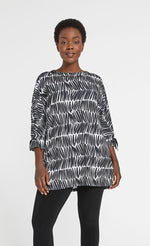 Load image into Gallery viewer, Front top half view of a woman wearing black leggings and the sympli halo tie cuff tunic in black and white shifted stripes. This tunic has 3/4 length sleeves that are tied with ribbon at the hem. The top has a boxy shape and sits below the hips.
