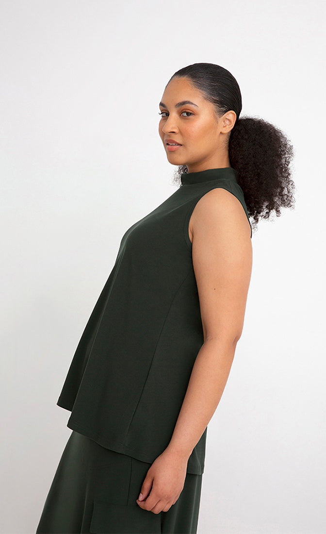 Left side top half view of a woman wearing the Sympli Mock Neck Tank in the color seaweed. This top is dark green, sleeveless, and features a mock neck.
