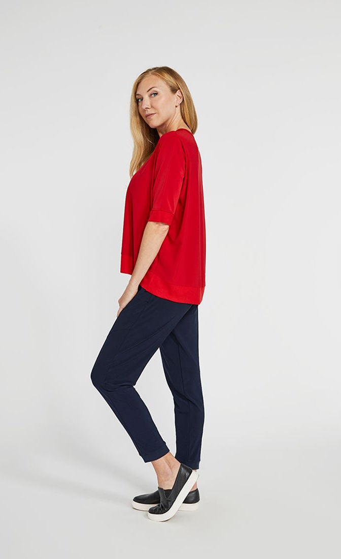 left side full body view of a woman wearing the sympli motion boxy top in the color poppy with blue bottoms. The top has 3/4 length sleeves and an asymmetrical double side-stepped hem.