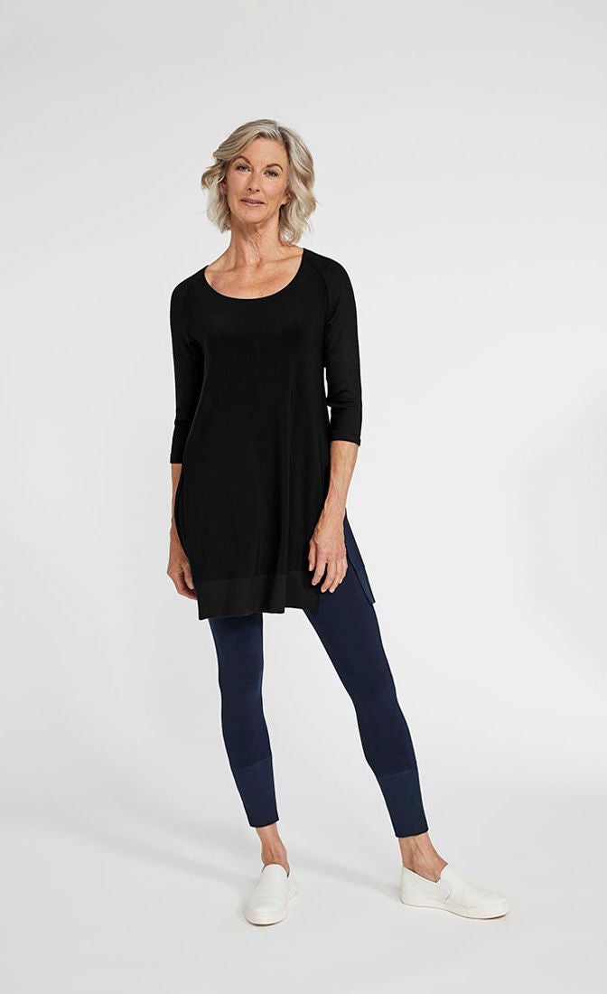 Front full body view of a woman wearing blue leggings and the sympli motion trim raglan tunic in black. This tunic has a scoop neck, 3/4 length ribbed raglan sleeves, a ribbed hem, and long side slits.