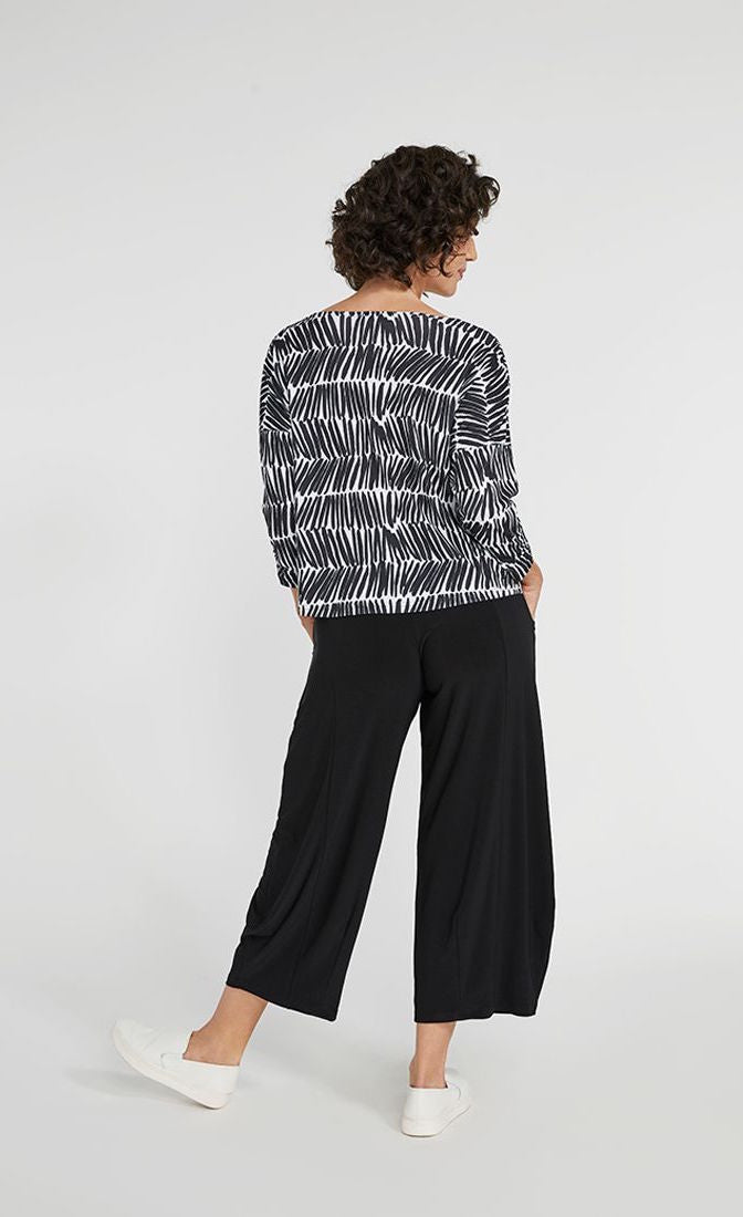 Back full body view of a woman wearing black pants and the Sympli Spark Boxy Top. This top has black and white shifted stripes. It has 3/4 length sleeves and sits slightly above the hips.