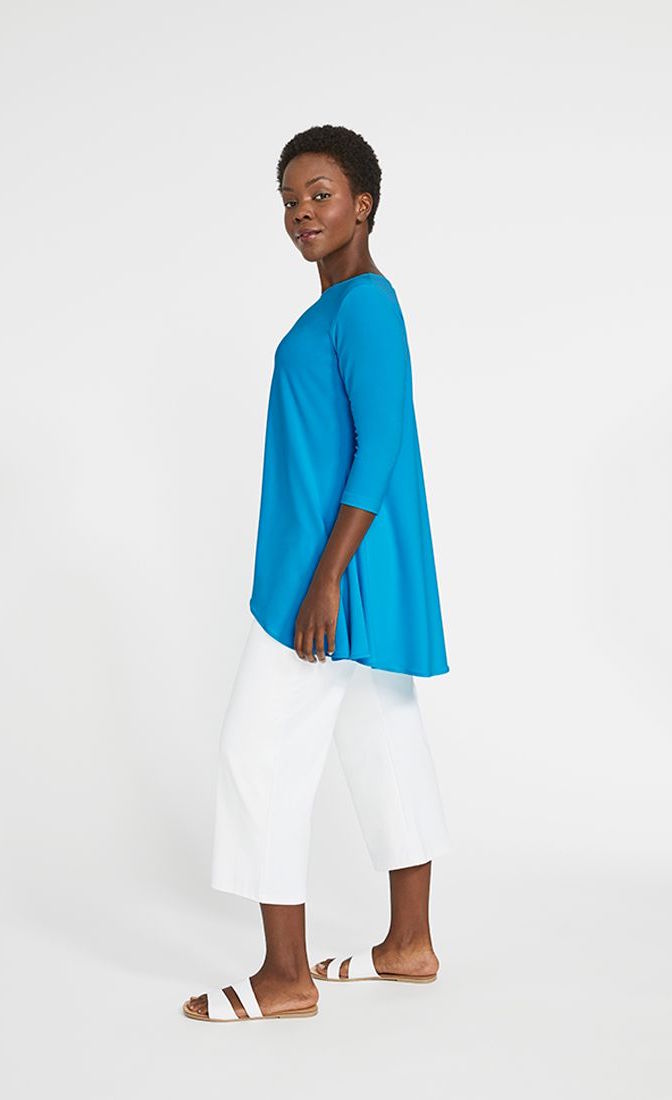 Left side full body view of a woman wearing the sympli true t in the color splash. This color is a sky blue. The top has 3/4 length sleeves, a boat neck, and a flowy high-low hem. On the bottom she is wearing white pants.