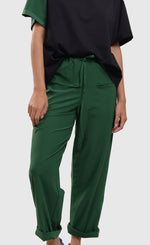 Load image into Gallery viewer, Front bottom half view of a woman wearing a black t-shirt and the alembika tekbika green drawstring pants. These pants are rolled up at the bottom and have two front pockets. The pant also has a drawstring waistband and a straight silhouette.
