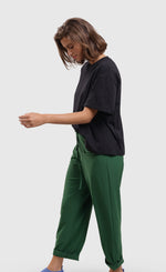 Load image into Gallery viewer, Left side full body view of a woman wearing a black t-shirt and the alembika tekbika green drawstring pants. These pants are rolled up at the bottom and have two front pockets. The pant also has a drawstring waistband and a straight silhouette.
