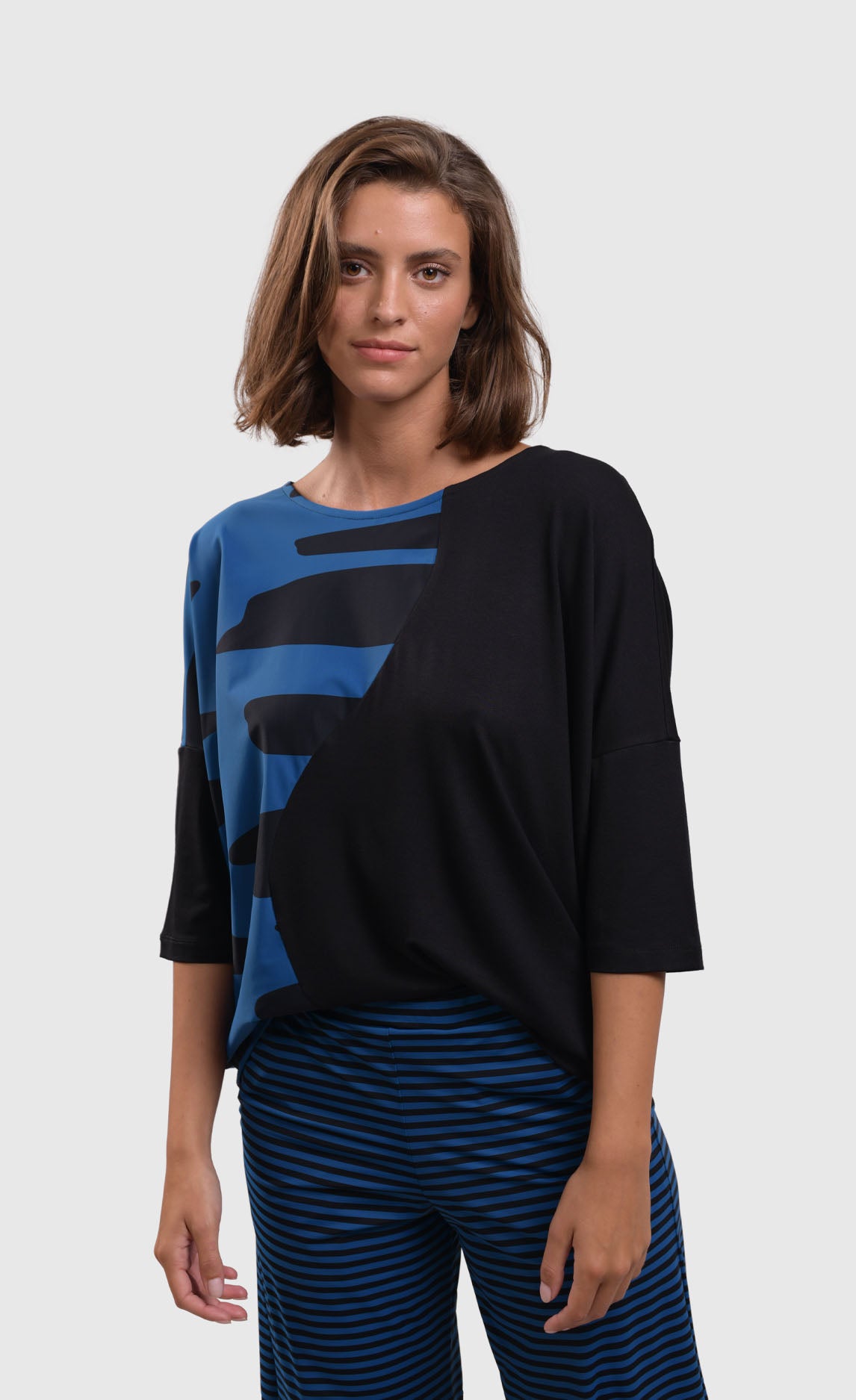 Front top half view of a woman wearing blue and black striped pants and the alembika tekbika ocean wave top. This top is black with a right sides wave paneling design in blue. The top as a scoop neck and drop shoulder, 3/4 length sleeves.
