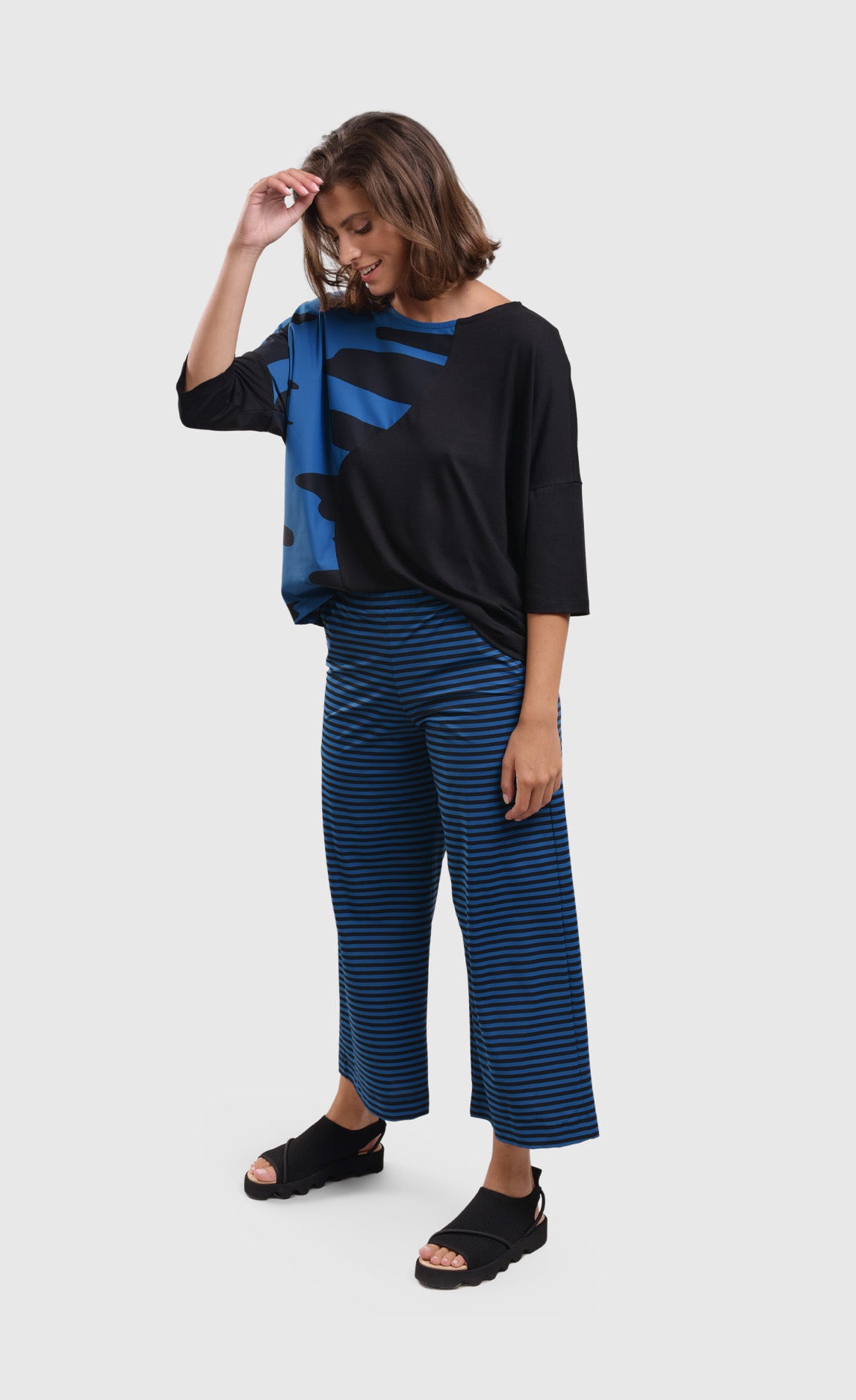 Front full body view of a woman wearing blue and black striped pants and the alembika tekbika ocean wave top. This top is black with a right sides wave paneling design in blue. The top as a scoop neck and drop shoulder, 3/4 length sleeves.