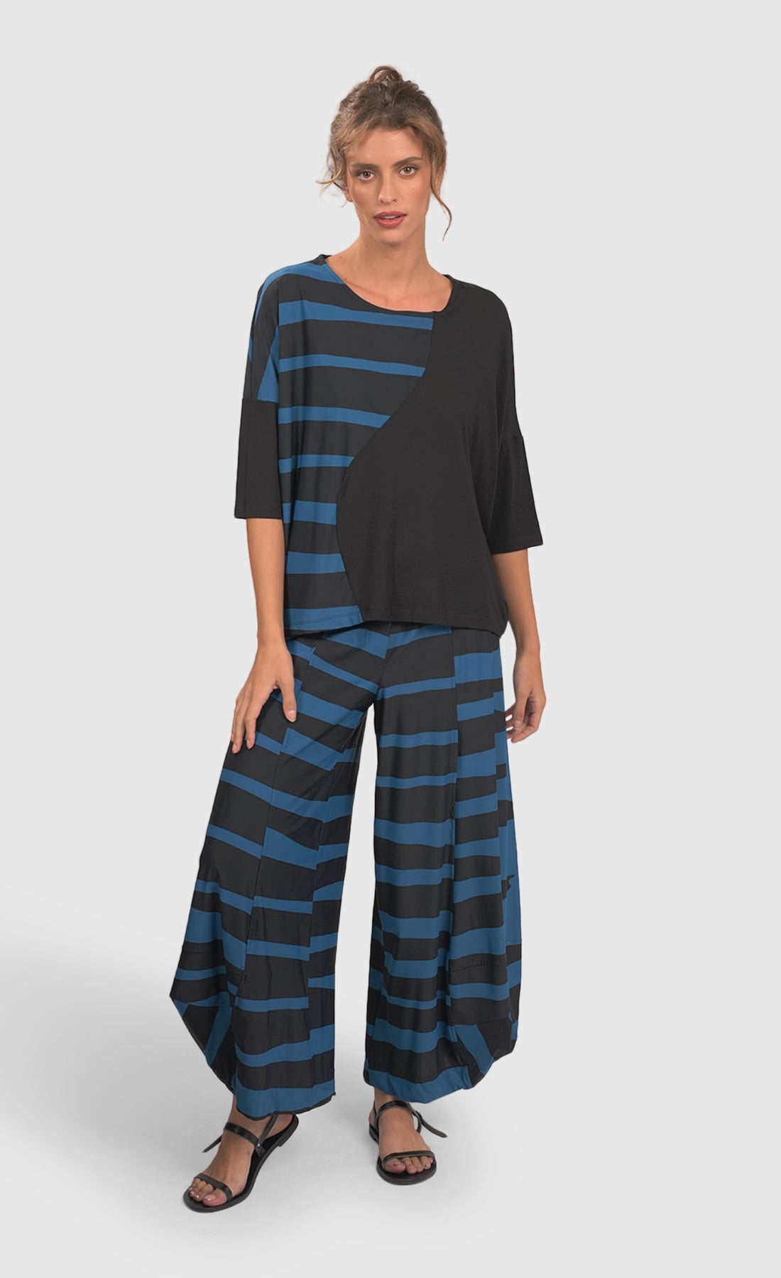 Front full body view of a woman wearing blue and black striped pants and the alembika tekbika ocean wave top. This top is black with a right sides wave paneling design in blue. The top as a scoop neck and drop shoulder, 3/4 length sleeves.