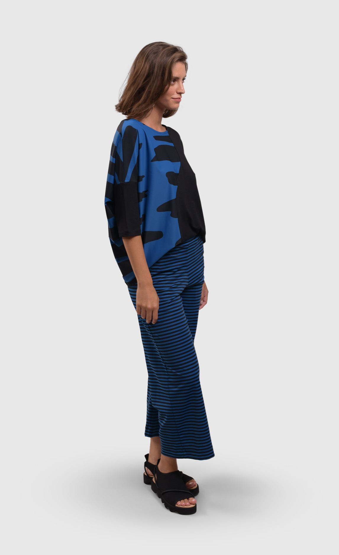 Front right side full body view of a woman wearing blue and black striped pants and the alembika tekbika ocean wave top. This top is black with a right sides wave paneling design in blue. The top as a scoop neck and drop shoulder, 3/4 length sleeves.