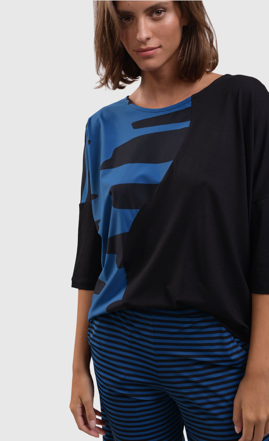 Front close up view of a woman wearing blue and black striped pants and the alembika tekbika ocean wave top. This top is black with a right sides wave paneling design in blue. The top as a scoop neck and drop shoulder, 3/4 length sleeves.
