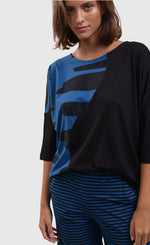 Load image into Gallery viewer, Front close up view of a woman wearing blue and black striped pants and the alembika tekbika ocean wave top. This top is black with a right sides wave paneling design in blue. The top as a scoop neck and drop shoulder, 3/4 length sleeves.
