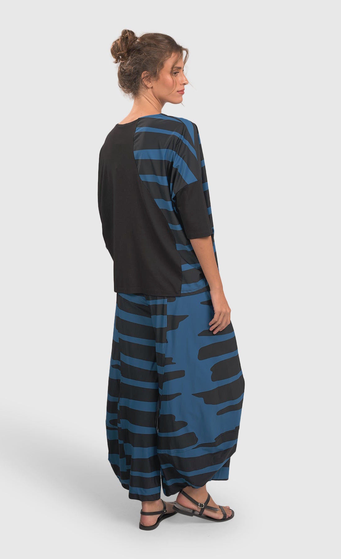 Back full body view of a woman wearing blue and black striped pants and the alembika tekbika ocean wave top. This top is black with a right sides wave paneling design in blue. The top also has drop shoulder, 3/4 length sleeves.