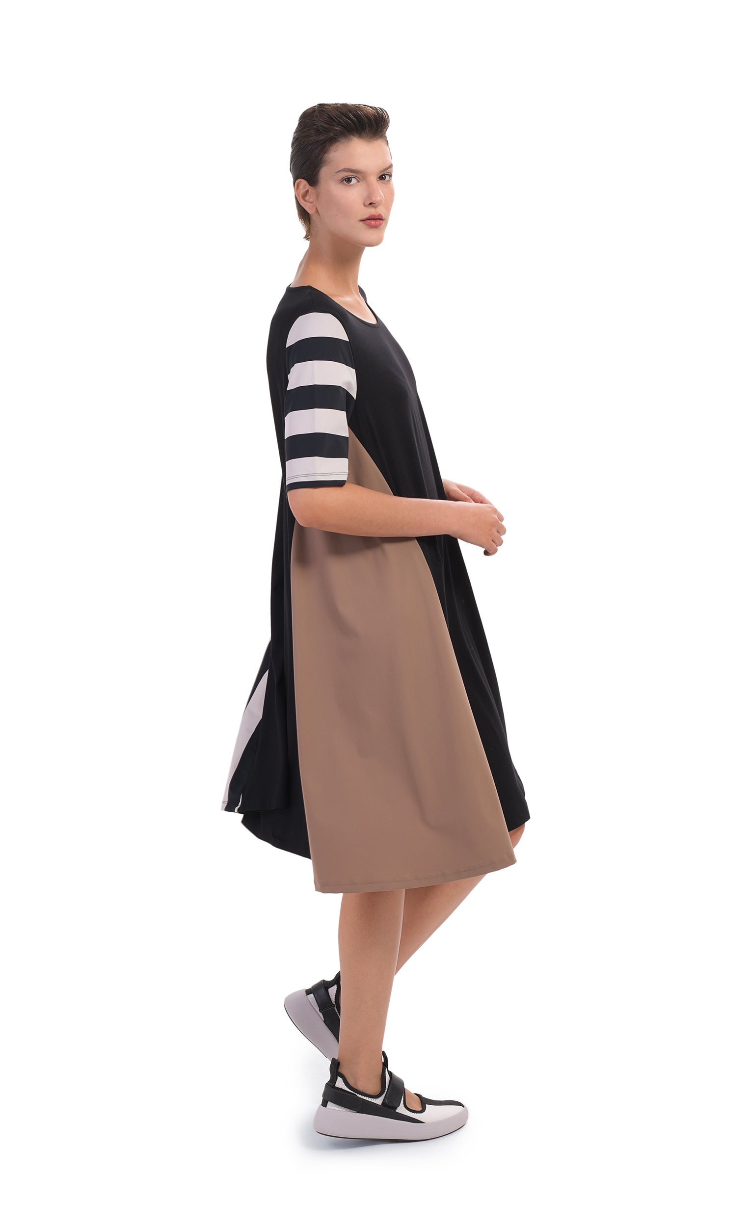 Right side, full body view of a woman wearing the alembika tekbika swing dress. This dress is black with one brown side and striped elbow length sleeves. The dress sits at the knees.