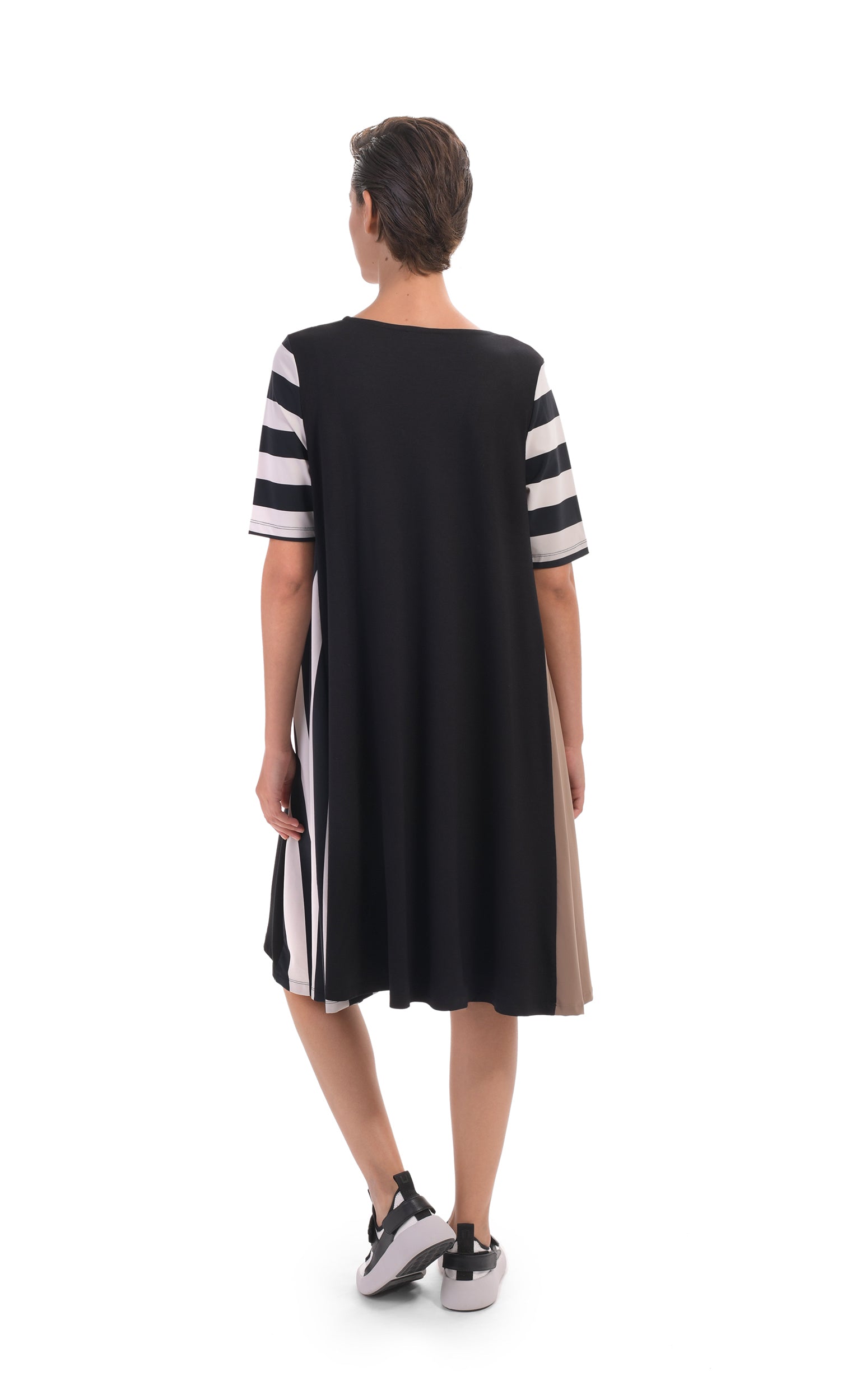 Back, full body view of a woman wearing the alembika tekbika stripe dress. This dress is black with one brown side, one striped side, and striped elbow length sleeves. The dress sits at the knees.