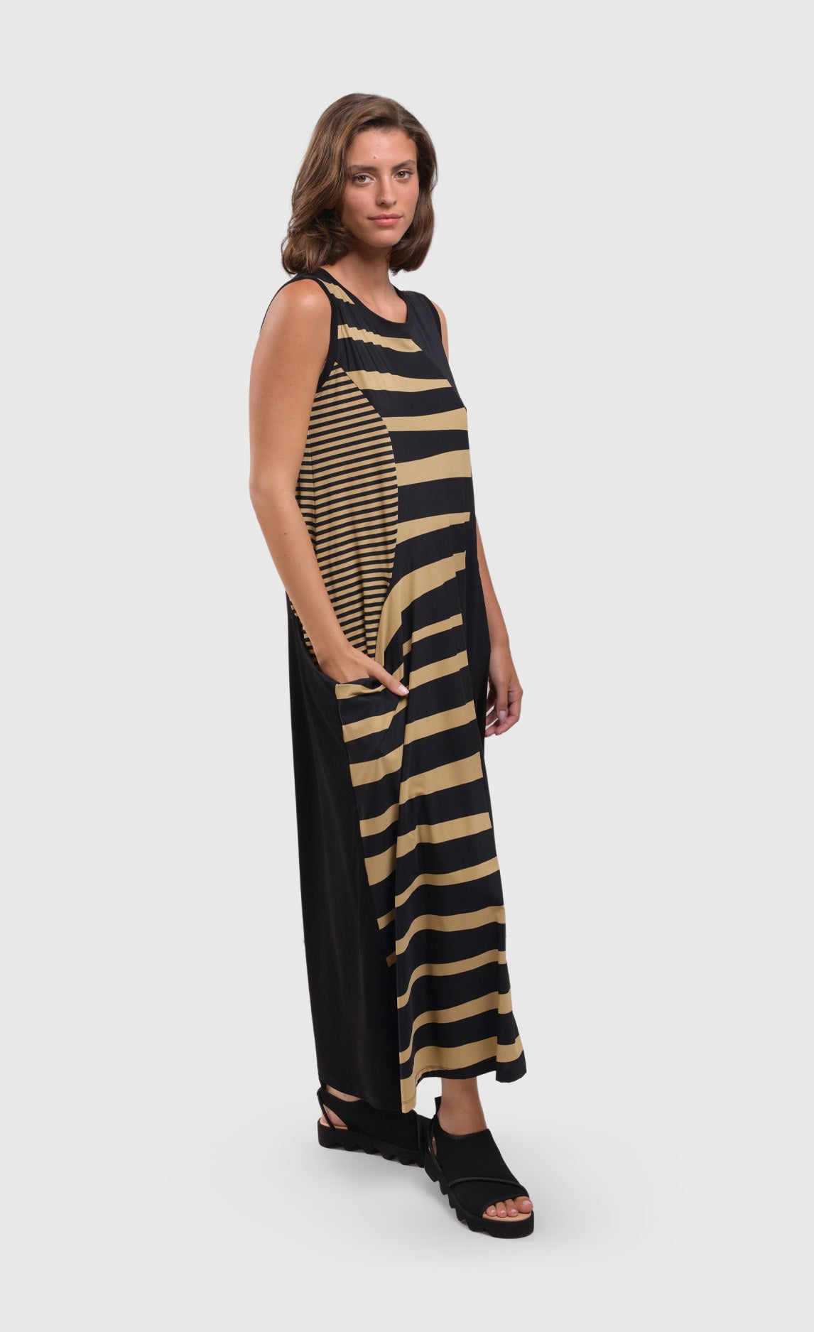 Front right side full body view of a woman wearing the alembika tekbika dunes wave maxi dress. This sleeveless dress is black with a wavy striped black and yellow print on the right side. The dress also has a single front draped pocket on the right side. 