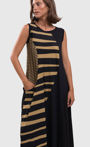 Front close up view of a woman wearing the alembika tekbika dunes wave maxi dress. This sleeveless dress is black with a wavy striped black and yellow print on the right side. The dress also has a single front draped pocket on the right side. 