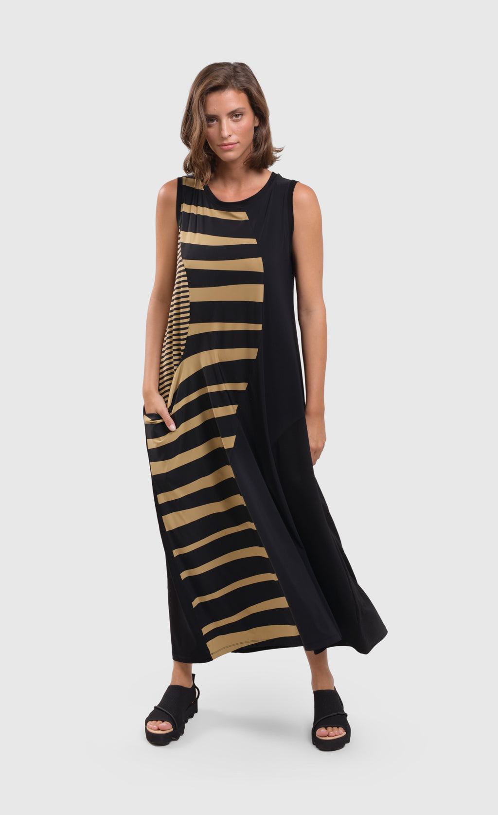 Front full body view of a woman wearing the alembika tekbika dunes wave maxi dress. This sleeveless dress is black with a wavy striped black and yellow print on the right side. The dress also has a single front draped pocket on the right side. 