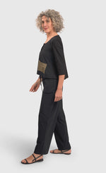 Load image into Gallery viewer, Left side full body view of a woman wearing black pants and the alembika tekbika dune crop jacket. This jacket is black with a single left sided front pocket that is striped black and yellow. The jacket has 3/4 length sleeves and a button down front.
