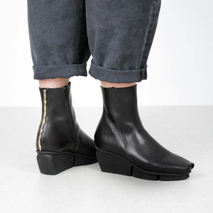 Outer side and back view of a woman wearing the trippen flaw boot in black waw. The back of this squared-toe boot has a cracked gold seam. The boot has a black wedged sole.