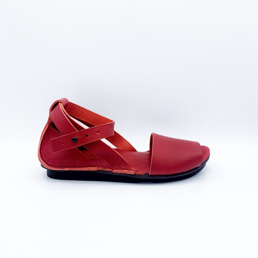 Outer view of the trippen iris sandal in red. This flat sandal has a wide strap over the toes, leather that covers the heel, and an ankle strap that criss crosses and attaches to the sole on both sides of the shoe.