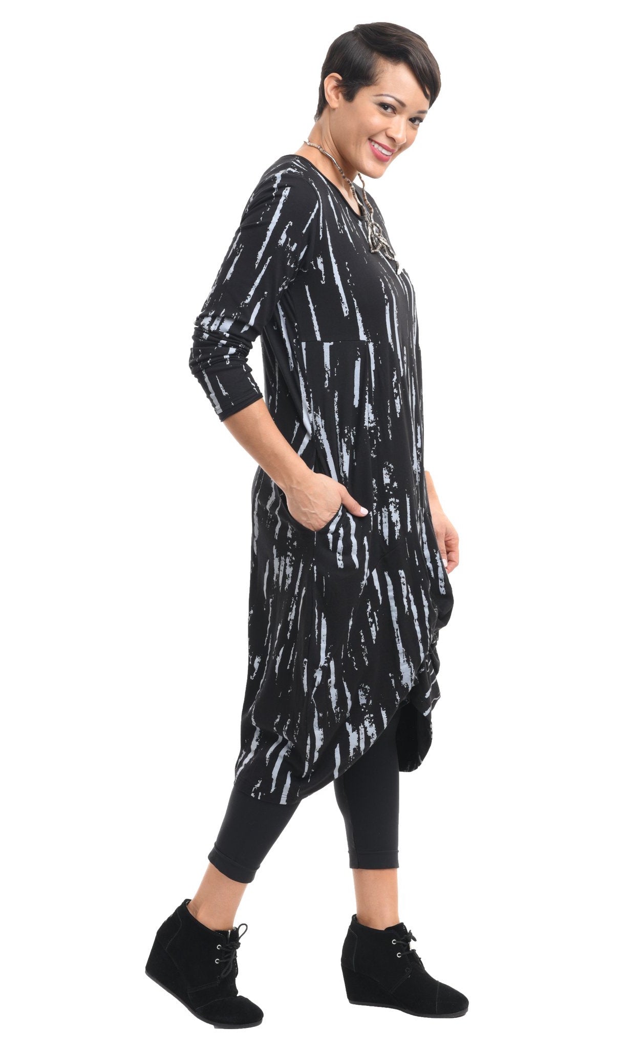 Right side full body view of a woman wearing the tulip karma dress in black with a grey airbrush print. The dress has long sleeves, a gathered knot in the front, and pockets.