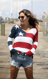 Front full body view of a woman standing on a boardwalk wearing the wooden ships flag v cotton sweater. This sweatter has the flag on it with a v-neck and long sleeves.