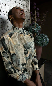 Front view of a woman laughing and wearing the unbreakable evolution petra blouse. This blouse is cream colored with a magazine cut out print all over it. It has long sleeves and a high neck.