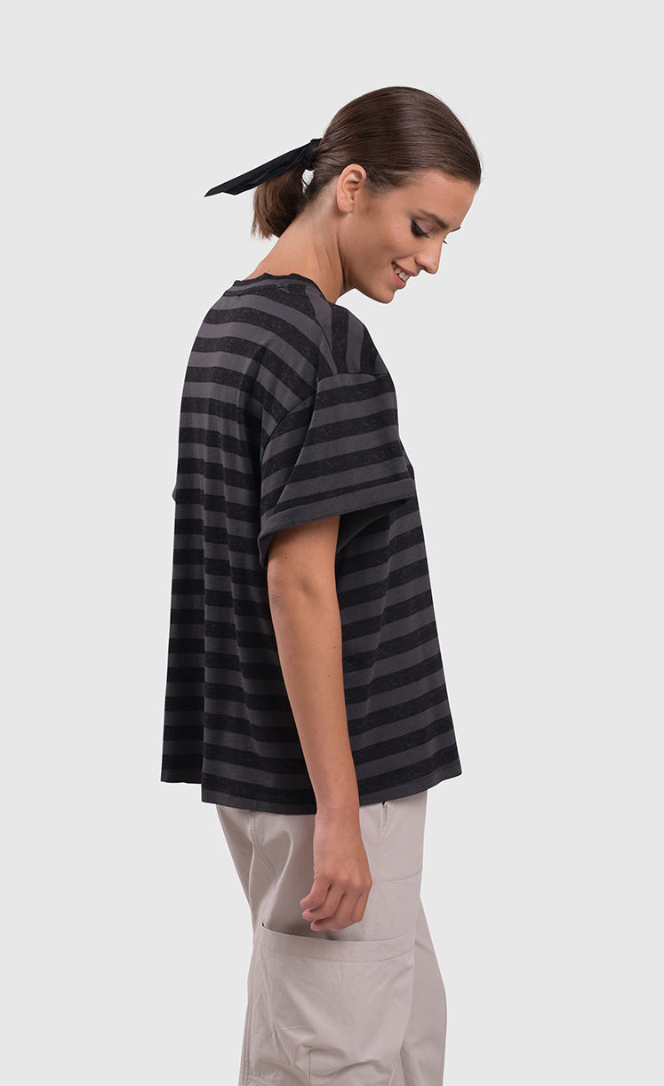 Right side top half view of a woman wearing the alembika urban stripe moon boxy tee. This top has grey and black striping, a crew neck, a wide silhouette, and short sleeves.