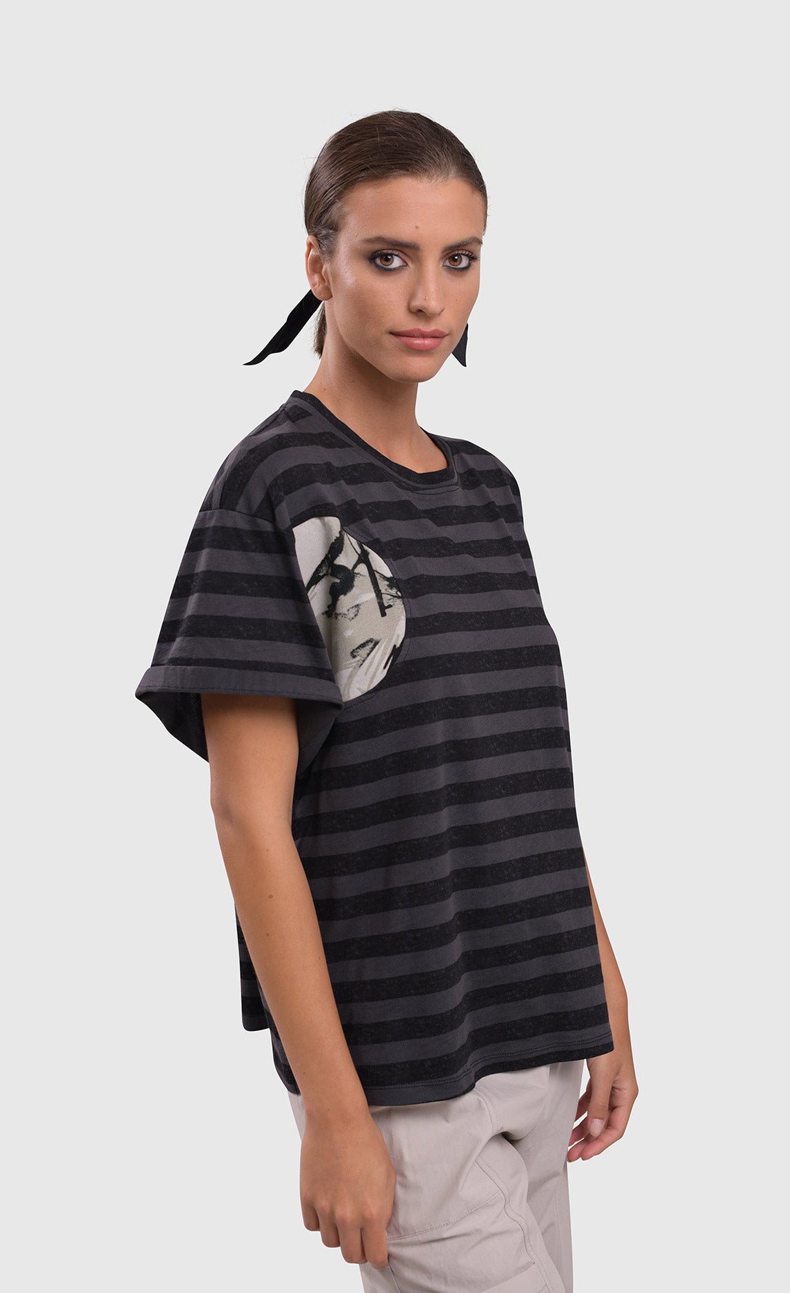 Front top half view of a woman wearing the alembika urban stripe moon boxy tee. This top has grey and black striping with two circular patches of a white fabric that have a scribble print on them. The top has a crew neck, a wide silhouette, and short sleeves.