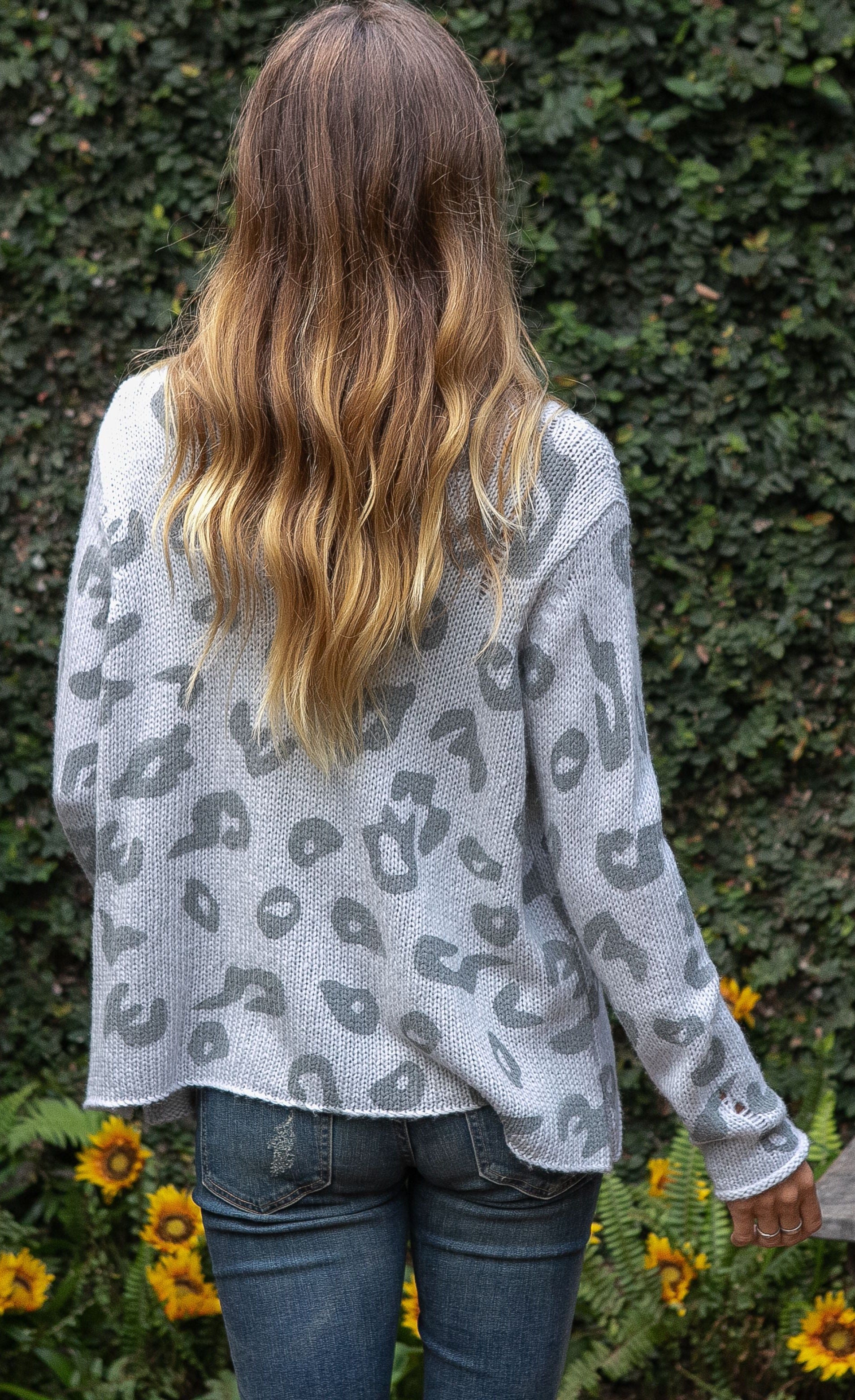 Back top half view of a woman wearing jeans and the wooden ships distressed leopard sweater. This sweater is grey with dark grey leopard print on the front. The is also distressed.