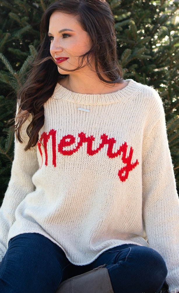 Front top half view of a woman sitting and wearing jeans and the wooden ship merry chunky crew sweater in the color alabaster/ginger. This sweater is white with the word merry written in red cursive on the front.
