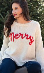 Load image into Gallery viewer, Front top half view of a woman sitting and wearing jeans and the wooden ship merry chunky crew sweater in the color alabaster/ginger. This sweater is white with the word merry written in red cursive on the front.

