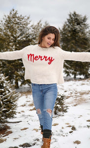 Front full body view of a woman walking through snow wearing jeans and the wooden ship merry chunky crew sweater in the color alabaster/ginger. This sweater is white with the word merry written in red cursive on the front.