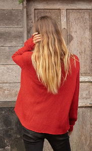 Back top half view of a woman wearing jeans and the wooden ships mini reindeer crew sweater. This sweater is poppy red colored with long sleeves.