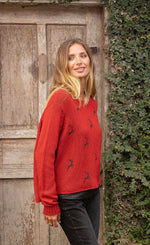 Load image into Gallery viewer, Front right side top half view of a woman wearing jeans and the wooden ships mini reindeer crew sweater. This sweater is poppy colored with a mini reindeer print on the front in the color ink. This sweater has long sleeves and a crew neck.
