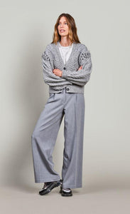Front full body view of a woman wearing grey pants and the summum balloon sleeve cardigan in the color fog/grey. This sweater has a cable knit pattern, a button down front, and fringe on the balloon sleeves.