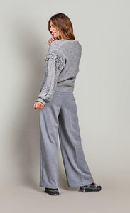 Back full body view of a woman wearing grey pants and the summum balloon sleeve cardigan in the color fog/grey. This sweater has a cable knit pattern, a button down front, and fringe on the balloon sleeves.